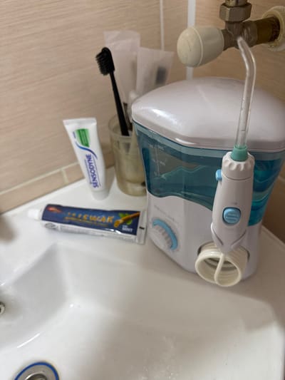 The image depicts a close-up view of a bathroom counter area, likely in a residence in Ciorescu, Moldova, though the specific location within the city cannot be independently verified from the image.
The scene focuses on dental hygiene items arranged on a white ceramic sink surface and against a light beige tiled wall. In the center-right, a white and translucent blue water flosser unit, branded "REVLYLINEA," stands upright. Its translucent blue reservoir displays measurement markings, and a light blue dial is visible on its front. A coiled white hose extends from the unit, connecting to a white handle with light blue buttons. A clear plastic flosser tip is inserted into the handle, which is further connected via a clear tube to a brass-colored pipe fitting on the wall with a white spherical valve.
To the left of the water flosser, a tube of "MISWAK" toothpaste, predominantly blue with "MINT" visible, lies horizontally on the sink surface. Further back and to the left, a white tube of "SENSODYNE" toothpaste, with green and blue accents, stands upright. Behind the toothpaste tubes, a clear glass or plastic cup contains several dental items, including a black toothbrush with its bristles pointing upwards and several other items wrapped in white paper sleeves. The wall consists of light beige rectangular tiles. The white sink basin is partially visible in the lower-left, showing a chrome drain with a blue accent. No individuals are present in the image, and no active interactions or ongoing actions are depicted.