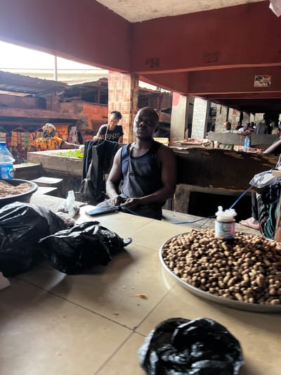 In Ọnịchạ, Nigeria, within a bustling marketplace stall, a man wearing a dark blue apron is seated behind a wooden counter. In front of him, a large platter of peanuts sits prominently, with a small, cylindrical bottle of what appears to be seasoning placed atop the pile. To his left, a black plastic bag lies on the counter, partially obscuring a mobile phone. Behind him, a woman in a vibrant, multi-colored dress and headwrap is visible, tending to green produce. Further in the background, other market-goers and stalls can be seen, suggesting a lively commercial environment. The stall is covered by a corrugated iron roof, and a red painted beam runs overhead, marked with the numbers "24" and "23".