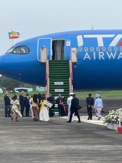 This image depicts a formal arrival or departure ceremony on an airport tarmac in Malabo, Equatorial Guinea. A large, blue ITA Airways passenger jet, clearly branded with "ITA AIRWAYS" in white lettering on its side and the "ITA" logo near the door, is the central object. A green mobile airstairs is positioned at an open door of the aircraft, facilitating access.

A diverse group of individuals is gathered on the tarmac, suggesting an official delegation or welcoming party. These include men in dark suits, several women dressed in formal attire, some holding bouquets of white flowers, and individuals in traditional or culturally significant clothing, such as a woman in a distinctive light blue and white patterned dress and another in a white dress with a yellow sash. Prominently featured is a man in clerical vestments, specifically a black cassock with a pink sash or stole, indicative of a bishop or cardinal.

A small flag displaying the yellow and white colors of the Vatican City, along with other colors, is mounted near the cockpit, strongly suggesting a papal visit or an event involving the Holy See. White floral arrangements are placed on a red stand near the base of the airstairs, adding to the ceremonial atmosphere. The sky appears overcast, indicating a cloudy daytime setting. Ground support equipment with visible text like "KING" and "CASA" is also present near the aircraft door. The overall scene conveys a sense of formal reception or farewell for an important dignitary.