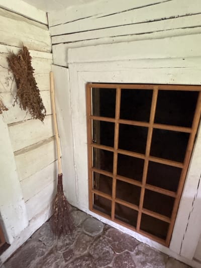 The image depicts a close-up view of a rustic, indoor scene, likely a corner of a room or attic. The primary subjects are a small, handmade broom and a wooden-framed window. The broom, lying on a white wooden floor, has a bundle of dried twigs or straw for bristles attached to a light-colored wooden handle. The floorboards are wide and show signs of age and wear, with visible seams and a slightly uneven surface, all painted white.

To the left of the broom, the wall is constructed of vertical white-painted wooden planks. To the right of the window, a rough, stone wall with irregularly shaped, grey and reddish-brown stones is visible, suggesting a very old or rustic construction.

The window is a key feature, with a dark, unreflective pane divided into a grid of 12 rectangular sections by a light-colored wooden frame. The window is set into a white-painted structure that appears to be part of the wall and ceiling. The overall impression is one of a simple, perhaps historical or miniature, interior space.

There is no discernible activity or event taking place; the scene is static. Notable details include the texture of the broom bristles and the stone wall, the planked construction of the wooden elements, and the grid pattern of the window. The lighting suggests an indoor setting, possibly with natural light coming from the window, though the interior is predominantly dark. There is no visible text in the image. The location context of Sielachowskie, Poland, adds a geographical element to the scene.