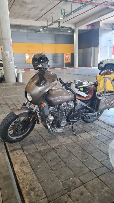 A metallic dark brown Indian brand motorcycle is parked on a surface of grey rectangular pavers within a covered structure, likely a parking garage or loading bay, in the city of Yokneam Illit, Israel. The motorcycle, positioned with its kickstand deployed, features a small front fairing, a brown solo seat, and dark hard-shell saddlebags with decals on both sides. A large yellow waterproof dry bag is secured to the rear fender. A black full-face helmet rests on the front fairing, and a white full-face helmet with green accents is placed on the passenger seat area. In the background, a concrete pillar and a wall composed of horizontal yellow and grey panels are visible. A red and white striped curb marking a boundary line is on the ground, with a red promotional sign on a stand further back. The exposed concrete ceiling displays red fire suppression pipes, grey electrical conduits, and fluorescent light fixtures. A white SUV is partially visible on the far right. No individuals are present in the scene.