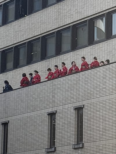The image displays a detailed view of a light-colored, multi-story building facade, likely in an urban setting. A group of approximately thirteen individuals, predominantly women, are lined up on a narrow architectural ledge or balcony spanning the width of the building. Most of the individuals are dressed in traditional Japanese kimonos, primarily in shades of red with intricate patterns, suggesting a cultural event or performance. One person on the far left wears a dark blue or black kimono with a lighter pattern. Their posture indicates they are standing and observing, with their attention directed outwards from the building. Above and below this group, rows of dark-framed rectangular windows are visible, reflecting elements of the surrounding environment. Given the traditional attire and the context provided, this scene is consistent with an observation point or public appearance in a historic district, such as the Higashi Chaya District in Kanazawa, Japan.