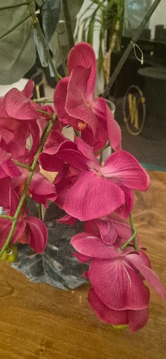 A close-up shot showcases artificial pink orchids. The petals exhibit a visible texture with slight color variations, adding depth. Stems connect the blooms, creating an arrangement, while the wooden floor serves as a contrasting backdrop. Other objects are slightly visible, enhancing the overall composition.