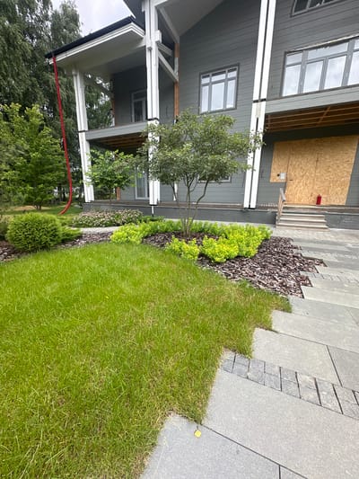 This image depicts an exterior view of a modern building with a landscaped garden in Uyut, Russia. The building features gray siding and several windows with dark frames. Parts of the building appear to be under construction or renovation, with plywood covering some areas. A red hose is visible, running across the lower portion of the frame.

The garden area includes a neatly mowed lawn, a decorative bed with dark bark mulch, and various plants, including a small tree with green leaves and some bright green shrubs. A pathway made of large gray tiles, interspersed with smaller gray stones, runs alongside the garden.

The scene suggests a daytime setting, with overcast or diffused lighting, hinting at possibly cloudy weather. The overall impression is one of a residential or commercial property with ongoing exterior work. There are no visible people or text in the image.