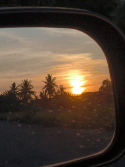 The image shows a sunset view seen through a car's side mirror. The setting sun is a bright orange orb, partially obscured by the horizon. Silhouetted against the bright sky are palm trees and a building with a dome, the foliage and structures indistinct. The mirror's frame is black and the view is slightly blurry. The scene is likely in or around the city of Cotbuket, Indonesia, based on the palm tree vegetation and architecture. The road visible in the lower part of the mirror is dark.