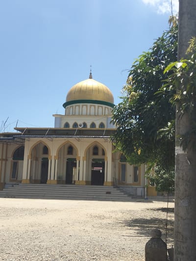 The image shows a mosque in Payademam Dua, Indonesia, under a clear blue sky. The mosque has a large golden dome with a green band around its base. The building is cream-colored with tan accents, featuring arched doorways and windows. A wide set of stairs leads up to the mosque's entrance. The foreground is a sandy or gravelly area. A leafy tree is on the right side of the frame, partially obscuring the mosque, and a concrete pillar is visible on the right edge.