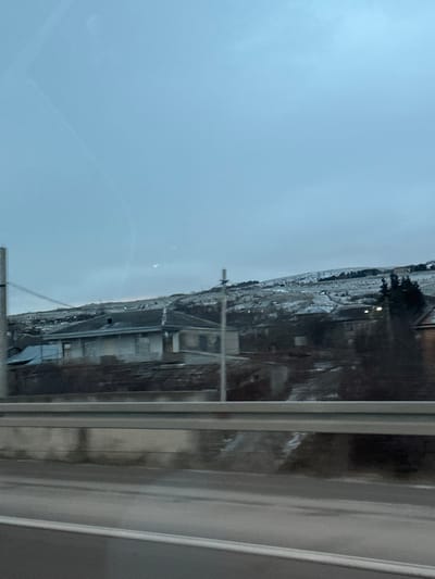 A view from a window shows a cityscape or townscape in the distance. The sky is overcast and grey. Buildings and terrain are visible, with some areas appearing to have snow cover. Reflections and smudges are present on the window. Window frames and part of a sill are in the foreground. The image is blurry and the details are difficult to discern.