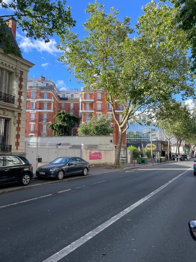 This image depicts a vibrant urban street scene in Malakoff, France, on a clear, sunny day. The sky is bright blue with scattered white clouds, and lush green trees with dense foliage line the street, casting shadows and providing shade.
On the left side of the street, architectural details vary. In the immediate foreground, a building with ornate classical design elements and a dark roof is visible. Behind it stands a taller, multi-story building featuring warm red brick facades, numerous windows, and small balconies. Further down, a modern building with a large glass facade reflects the surroundings, suggesting a commercial establishment. A yellow oval sign, possibly reading "Mias," is prominently displayed on the glass building, and a green cross sign, indicative of a pharmacy, is also visible. Outdoor seating with tables and green planters can be discerned in front of this area.
The right side of the image features a dark asphalt road with a distinct solid white line running down its length. Several dark-colored cars are parked parallel to the curb. One parked car clearly displays a French license plate with the registration "BC-444-DC." Along the sidewalk on the right, a pink sign is visible, reading "P Parking à louer" (Parking for rent). The overall scene portrays a typical, bustling urban environment during daylight hours.
