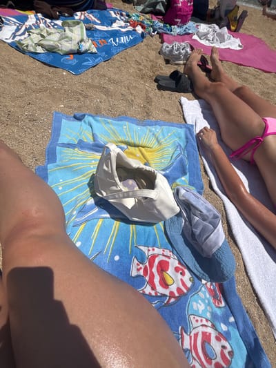 The image shows a sunny day at a beach in Nea Iraklitsa, Greece. Several towels are laid out on the sand, with various items of clothing and footwear scattered around. In the foreground, a person's tanned legs are visible, resting on a blue towel with a fish pattern. Nearby, there is a white bag, a pair of blue slippers, and a white towel. In the background, other people's legs and feet are visible, as well as more towels and beach accessories. The water and waves of the sea are visible in the upper right corner. The overall scene depicts a typical summer day at the beach.