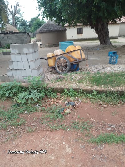The image depicts an outdoor scene in the city of Dokan Tofa, Nigeria. In the foreground, a stack of grey concrete blocks is visible. To the right of the blocks, a hand-pulled cart is laden with yellow plastic containers. A large tree with thick, gnarled branches dominates the background, partially obscuring a white building with a corrugated metal roof. Several traditional round huts with thatched roofs are also present, along with a blue plastic crate. The ground is a mix of dirt, gravel, and sparse vegetation. The "Samsung Galaxy A03" watermark is present in the bottom left corner, indicating it's a photograph taken with that device.