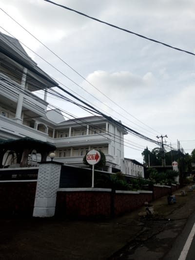 A white, multi-story building with balconies sits behind a brick wall. An "OUT" sign with a right-pointing arrow stands near the wall. Power lines crisscross the sky above the building. The scene is overcast, and a glimpse of a road suggests the view is taken from across the street. Additional signage and greenery are visible further down the wall. A utility pole stands on the right, against a cloudy sky.