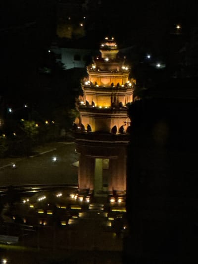 The image shows a tall, ornate tower structure illuminated at night. The tower has multiple tiers and is decorated with numerous small, bright lights. The structure appears to be made of stone or concrete and has arched openings and decorative elements. In the foreground, there is a dark, blurry mass that obstructs a portion of the view. The background is dark, with some indistinct shapes and scattered lights suggesting the presence of other buildings or distant streetlights. The ground in front of the tower is paved and reflects some of the ambient light. The overall scene suggests a nighttime view of a monument or building in a city setting. Given the location specified as Khan Boeng Keng Kang, Cambodia, this structure is likely the Independence Monument in Phnom Penh, a well-known landmark in that district. There are no discernible people or specific actions depicted.