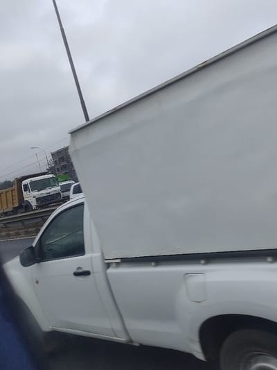 A white pickup truck with a large, boxy, white canopy is driving on a road. The canopy almost completely obscures the cab of the truck. Other vehicles are visible in the background, including a tan dump truck and a white truck with a green side. An overhead electrical pole is prominent in the background against a cloudy sky. The image appears to be taken from inside another vehicle, possibly moving along the same road.