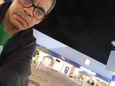 The image depicts an outdoor commercial scene at night in Tumbaco, Ecuador. In the upper-left foreground, a male subject with short, graying hair, dark-rimmed glasses, a green shirt, and a black puffer jacket is visible, looking directly towards the camera. The background features a paved plaza with an outdoor seating arrangement comprising multiple white square tables and white chairs, shaded by several open green and white striped patio umbrellas. A building with a light blue and white facade extends across the mid-ground, displaying an illuminated orange rectangular logo and a partial white sign with blue text. Several indistinct individuals are present in the background, standing or walking near the outdoor seating and building entrance. The dark sky indicates nighttime, with the area artificially illuminated.