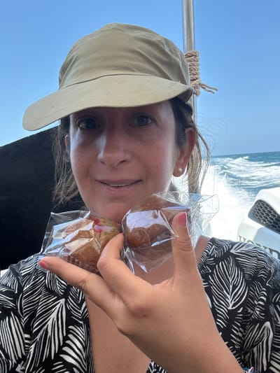 An image taken in Pak Nam, Thailand, depicts a female individual on a moving boat under a clear blue sky. The woman, who appears to be of middle age, wears a light tan bucket hat and a dark garment with a light leaf pattern. She exhibits a slight smile, revealing her teeth, and has a single white pearl earring visible in her right ear. Her left hand, with light pink painted fingernails, holds two individually plastic-wrapped muffins prominently in the foreground. One muffin, positioned to the left, appears to contain red fruit pieces, while the other is a plain brown variety. In the background, turbulent water and white wake are visible on the right, indicative of a vessel in motion. A white outboard motor bearing the "HONDA" brand logo is partially visible on the far right. A vertical metal pole wrapped with light-colored rope is positioned to the right of the woman's head, suggesting boat infrastructure.