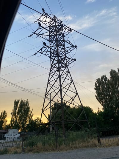 The image shows a tall, lattice-style electricity pylon against a sky that transitions from blue with wispy clouds to a soft yellow-orange, suggesting either sunrise or sunset. Several power lines extend from the pylon. The pylon is dark brown or black and appears to be made of metal. It is positioned to the left of the frame, with its base obscured by the bottom edge of the image.

To the right of the pylon, there is a line of trees with dense green foliage. Some of the trees are tall and slender, while others are more bushy. Behind the trees and the pylon, there is a faint outline of a fence. In the lower right portion of the image, a paved surface, likely a road, is visible.

A white sign with black Cyrillic text is attached to the fence. The visible text reads "СТОЯНКА ИНОК". This likely translates to "Parking for monks" or "Monks' parking."

The overall scene conveys a sense of rural or semi-rural industrial infrastructure combined with nature. The lighting suggests a calm and peaceful time of day. There are no people visible in the image. The setting appears to be outdoors, possibly on the outskirts of a settlement given the presence of the electricity pylon and the road.
