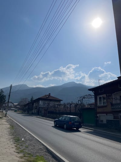 The image displays a street scene in Resilovo, Bulgaria, under bright daylight conditions. The sky is a clear, light blue with several white cumulus clouds scattered across the mid-horizon. The sun is prominently visible in the upper right quadrant, appearing as a bright white orb and causing a lens flare effect. A range of dark blue-grey mountains extends across the background, rising high above the settlement and appearing hazy in the distance.
An asphalt road runs from the lower left, gently curving uphill towards the right side of the frame. A paved sidewalk or verge, composed of light-colored interlocking pavers, borders the left side of the road. A wooden utility pole is positioned on the left, with approximately seven parallel overhead electrical wires stretching diagonally upwards and to the right across the sky.
Several buildings, primarily residential, line both sides of the road. On the immediate right, a two-story house is visible, featuring a brown tiled roof, dark wooden cladding on its upper floor, and lighter-colored walls on the lower floor, with multiple windows. A bare trellis structure, likely for climbing plants, extends from its facade, and a small balcony is visible on the upper right. Adjacent to this house on the right is a structure with a dark green garage door or gate. Further down the road on the left, additional houses with traditional red-tiled roofs are partially visible amidst sparse trees, some with bare branches.
A dark blue station wagon, identifiable by its Bulgarian license plate (partially legible as B 0320 XM), is driving on the asphalt road, moving away from the viewer towards the right side of the frame. No individuals are visible within the scene.