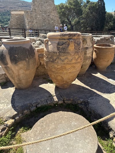 The image captures an outdoor archaeological site, likely Knossos Palace in Heraklion, Crete, Greece, under a bright, sunny sky. In the foreground, several large, ancient clay amphorae are prominently displayed. The amphorae vary in size and decoration; one is particularly large and decorated with intricate geometric patterns and stylized motifs. To the left, another large amphora is visible, showing signs of age and wear. Further back, smaller amphorae and pottery fragments are scattered amongst the ruins.
Behind the amphorae, the remains of stone structures, characteristic of Minoan architecture, form the backdrop. Tourists, identifiable by their casual attire and the presence of a guide holding an umbrella, are seen observing the artifacts from a raised walkway behind a wooden railing. The environment is characterized by rocky terrain, patches of green vegetation, and the harsh sunlight casting distinct shadows. The overall scene conveys a sense of historical significance and the ongoing preservation of ancient artifacts.