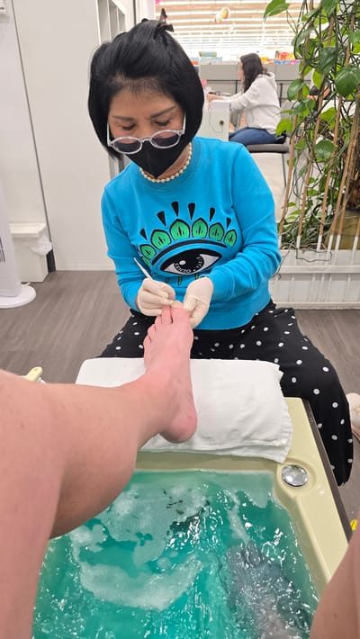 A person's foot is submerged in a bubbling, turquoise foot bath while a nail technician, wearing gloves and a mask, works on their toenails with a metal tool. The technician, with short black hair and glasses, is dressed in a blue sweatshirt with a large eye design and black polka dot pants. The scene appears to be a nail salon in Milton, Canada. In the background, another person wearing a white hoodie is visible at a counter. The environment includes a tiled floor, a white fan, and a large plant in a pot.
