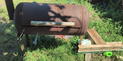 An old, rusty metal barrel barbecue, likely homemade, stands on metal legs in a grassy outdoor area. The barbecue has a visible handle made of light-colored wood and a chimney pipe extending upwards on the left side. Two small, looped metal pieces hang from the underside of the barbecue. In the background, there is dense green vegetation and some wooden planks forming a makeshift structure. A bright green plastic object is visible on the ground near the wooden planks. The scene suggests a rustic, possibly unkempt, backyard or outdoor space. The presence of a shadow of leaves on the barbecue indicates it is daytime with sunlight filtering through trees. The location is identified as being in the city of Есипово, Russia. There are no people or animals visible in the image.