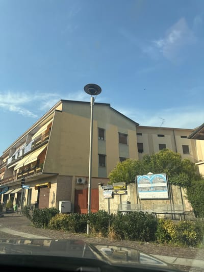 The image shows a street scene in Rudiano, Italy. The focus is on a multi-story building with a light yellow facade and balconies with awnings. A tall lamp post stands in front of the building. To the right of the building, there is a large billboard with Italian text, possibly an advertisement or event poster. Green bushes and a paved area with a cobblestone pattern are visible in the foreground, with the reflection of a car's dashboard on the lower part of the image. The sky is clear blue with a few wispy clouds. The overall impression is a sunny day in a European town.
