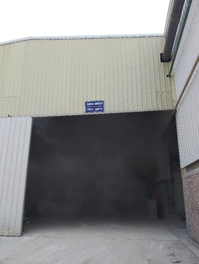The image shows the exterior of a building with a large open doorway. The doorway is located on the left side of the building and is partially obscured by a large metal door. The doorway is open and there is a large amount of dust and debris inside. The building has a corrugated metal exterior and the words "SODA DRYER" are printed on a blue sign above the doorway.  The building is located in the city of Pangasia, Bangladesh, at the coordinates (latitude: 22.693, longitude: 90.601).  There are no people or animals visible in the image. The building is located in a commercial or industrial area, with a concrete floor and visible brick walls.  There appears to be a black pipe protruding from the building wall on the right side of the image. There is also a red object that may be a fire extinguisher located in the interior of the building to the right of the doorway. There is no indication of the time of day when the image was taken.