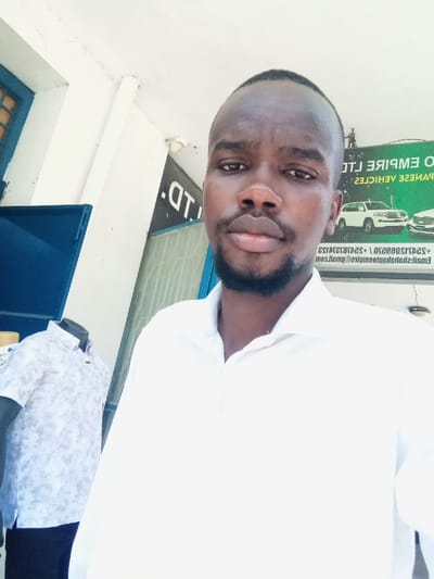 A daytime photograph shows a dark-skinned adult male with short dark hair and a short beard, wearing a white collared shirt, looking directly at the camera in a selfie-style composition. The scene is located in a commercial area, likely an outdoor or semi-enclosed space, within the Ganjoni/Shimanzi ward, Kenya. To the left of the subject, a headless and armless mannequin torso displays a light-patterned short-sleeved shirt over dark trousers. Behind the subject and to his right, a large dark sign with white, mirrored text is visible. Legible portions of the mirrored text include "O EMPIRE LTD", "PANESE VEHICLES" (indicating "JAPANESE VEHICLES"), phone numbers, and an email address "sales@GanjoniEmpire.com", identifying the establishment as Ganjoni Empire Ltd., a vehicle sales business. A partial blue window or door frame is visible on the left side of the frame. The overhead structure is a white ceiling with minor surface irregularities.