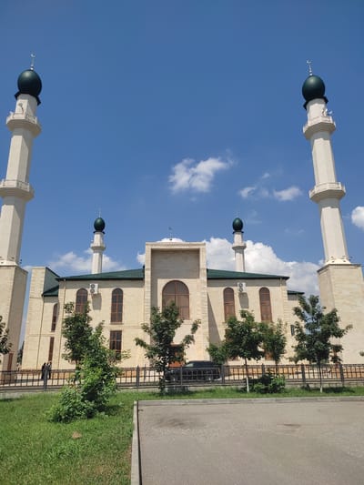 This media file depicts a mosque in Goyty, Russia, on a clear, sunny day. The image is oriented vertically, with the sky occupying the majority of the frame at the top. Several minarets, topped with green domes and crescent moons, rise towards the blue sky, which is dotted with fluffy white clouds. The mosque itself is constructed from light-colored stone and features arched windows with wooden frames. Green trees and manicured grass surround the building, and a black metal fence runs along the property line. A paved road is visible to the right of the mosque. The overall impression is one of peace and serenity. There is no visible text in the image.