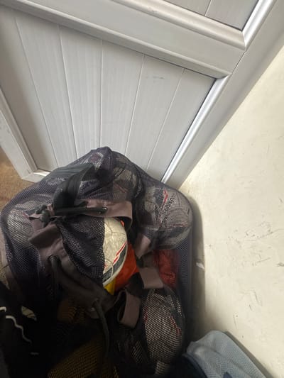 The image depicts a mesh bag filled with soccer balls and other items, positioned against a white wall and a white louvered door. The bag is dark and the contents are partially visible, including the patterned surfaces of the soccer balls. The environment appears to be indoors. The location of Charentsavan, Armenia is not verifiable based on visual information within the image.