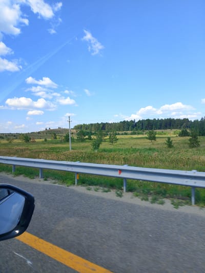 The image depicts a view from a moving vehicle on a highway. A metal guardrail runs along the side of the road, separating the highway from a grassy field dotted with trees. In the background, a gentle hill rises, covered in vegetation and a line of trees forming a dense forest. A utility pole stands on the hill. The sky is a bright blue with scattered white clouds. The overall scene is one of a rural, natural landscape under a clear sky. The location is identified as Алтынды ауылдық округі, Kazakhstan.