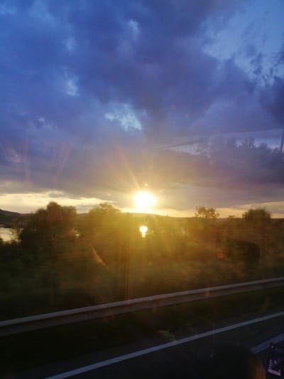 This is a photograph taken at sunset, possibly from inside a vehicle traveling on a road near Pocoleni, Romania. The sky is filled with dramatic, dark purple clouds, with the setting sun breaking through on the right side of the frame, casting a bright, golden light and visible sun rays. Below the sky is a landscape of trees and a body of water, likely a lake or river, reflecting the warm light. A guardrail runs horizontally across the lower portion of the image, suggesting the viewpoint is from a road. The image appears to have been taken through glass, as there are reflections visible on the surface, including what might be the interior of a vehicle and a person's head in the bottom right corner, holding a phone. The overall impression is one of a scenic, yet somewhat obscured, natural environment at dusk.