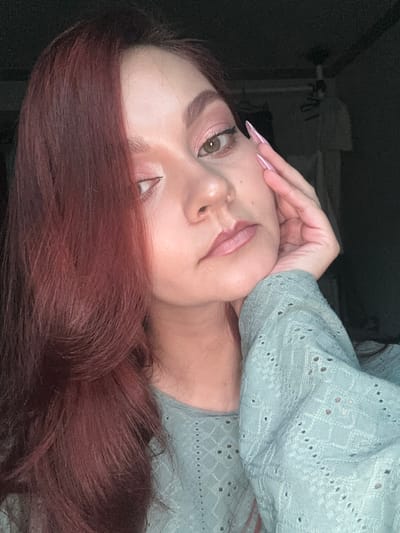 A young woman with long, auburn hair is posing for a portrait. She is wearing a light blue, textured sweater. Her nails are long and painted a light pink. She is touching her forehead with her hand. The background is dark and indistinct. There is no indication of Buk-myeon, South Korea.