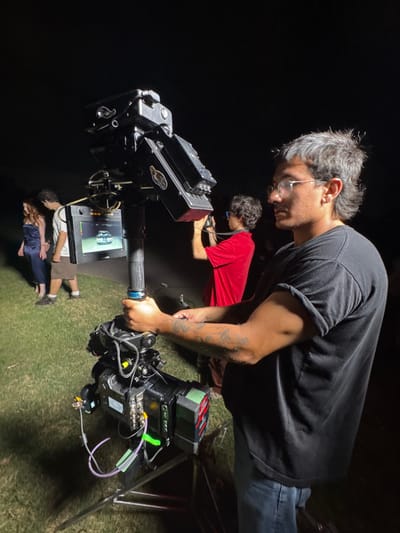 A camera operator, wearing glasses and a black shirt, uses a professional camera rig. He leans over, intently focused on the monitor displaying the scene being filmed. In the background, another person in a red shirt looks at a small screen, possibly assisting with the camera work. On the ground below, two people, presumably the actors, are positioned closely together in the darkness.