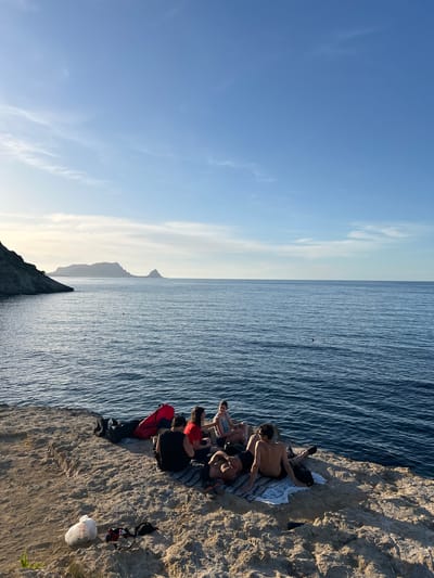 The image depicts a coastal scene in Sperone, Italy, at what appears to be late afternoon or early evening, characterized by warm, low-angle sunlight. The sky is predominantly clear blue, with scattered, wispy white clouds concentrated on the left horizon, which is lighter in tone. The sea, a calm deep blue, shows gentle ripples across its surface. In the mid-ground, a dark, indistinct object is visible in the water.
Across the distant horizon, a series of dark, silhouetted landmasses or mountains rise from the sea, with a more pronounced peak on the right. The foreground features a light-colored, textured rocky outcrop or cliff face, where a group of approximately seven individuals are gathered. The rock surface is uneven, showing shadows consistent with the sun's low position. Sparse green vegetation is visible on the far left edge of the outcrop.
The individuals are arranged in small clusters on blankets or towels spread on the rock. From left to right:
- A red duffel bag and a dark backpack are placed on the rock.
- A person in a black t-shirt sits with their back to the viewer, facing the sea.
- To their right, a person in a red t-shirt sits, also facing the sea.
- Further right, two individuals are relaxing on a striped blanket. One person (bare-chested) lies on their stomach, facing away from the camera. Another person (bare-chested male) sits upright with their back to the camera, wearing dark shorts and shoes.
- On the far right, two more individuals are seated. One person in a light top sits with their back to the camera, and another person with dark hair sits facing the sea.
- A white plastic bag, bearing the text "Todis," rests on the ground in the far left foreground, alongside scattered items of clothing and footwear. A dark, small object (possibly a speaker) is visible on the rock near the central group.
The individuals appear to be relaxing and conversing in a casual manner, enjoying the coastal environment.