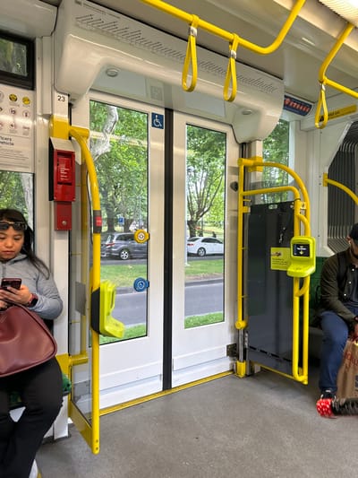 The interior of a public transit vehicle features yellow support structures and white panels. A person is seated near a closed door, while another passenger is engrossed in a phone. Outside, trees and parked vehicles are visible through the windows. A red emergency stop button is mounted near a window. The scene portrays an everyday moment on public transport.