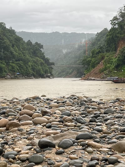 The image depicts a natural scene of a riverbank and the adjacent river. The foreground is dominated by a dense collection of smooth, rounded pebbles and stones of varying sizes and colors, ranging from light tan to dark gray. These stones are densely packed, forming a textured ground.

To the left of the river, a steep, vegetated hillside rises, covered in lush green trees and foliage. The hillside appears rugged and slopes down towards the river. In the middle ground, a wide, murky river flows. The water is a light brown or tan color, suggesting it might be carrying sediment.

Further into the background, on the left side of the river, a distant mountain range is visible, shrouded in mist and a cloudy sky. This indicates overcast or rainy weather. A striking orange construction crane is visible on one of the lower mountain slopes, suggesting ongoing development or infrastructure work in the area. A suspension bridge structure can also be faintly seen in the distance. On the right bank of the river, there are some indistinct structures and a few small boats or rafts are visible in the water.

The overall mood of the image is serene and natural, with a hint of human activity through the presence of the crane and boats. The weather appears to be overcast and potentially damp, contributing to the misty atmosphere of the distant mountains. The scene is likely in a rural or remote location, given the natural landscape and the presence of the river and mountains. No visible text is present in the image.