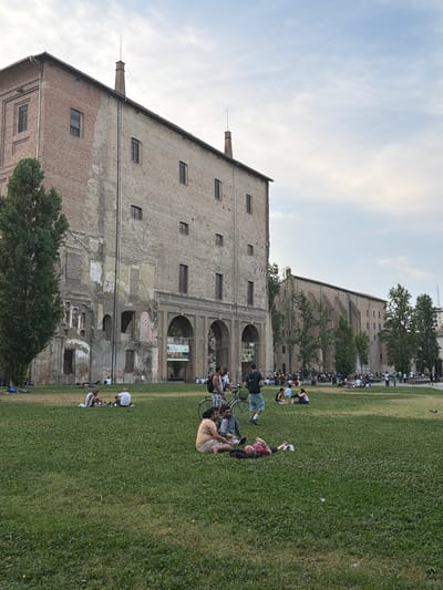 The image depicts an outdoor scene in Parma, Italy, likely during the late afternoon or early evening given the soft lighting and clear sky with some light clouds. A large, old brick building with multiple windows and archways dominates the left side of the frame. The building appears to be a historical structure, possibly a former industrial building or a large residence, with some signs of age and wear on its facade.
In front of the building, a large grassy field stretches out, where numerous people are gathered. Many are seated or lying on the grass, enjoying a leisurely day outdoors. Some are interacting with each other, while others appear to be relaxing or observing their surroundings. There are several bicycles visible, suggesting that people have arrived by bike. The atmosphere seems relaxed and communal, with individuals and small groups spread across the lawn.
Further back, more people are visible, forming a continuous line along the edge of the field, closer to another building. This area also appears to be part of the historical complex. The presence of many people enjoying the park suggests a popular public space, possibly a gathering for an event or simply a common recreational area. The overall impression is one of a pleasant, warm-weather day in a historical Italian city, with people taking advantage of the outdoor space. No visible text is present in the image.