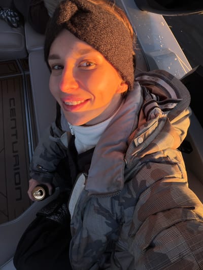 A woman wearing a dark beanie and a plaid hooded jacket sits inside a Centurion boat. She holds a gold-colored can in her gloved hand. The setting sun casts an orange glow on her face. The location is possibly near Терехово, Russia.