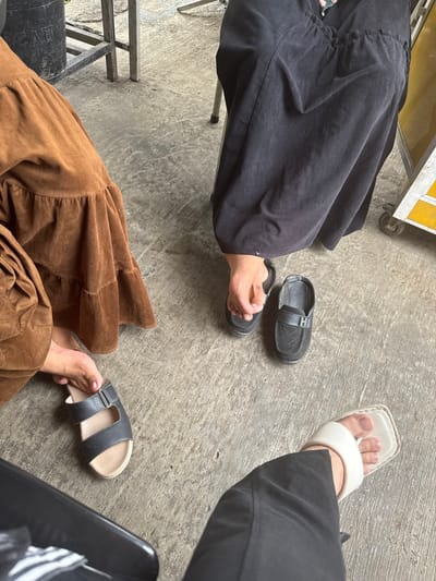 The image shows a close-up view of three people's feet and lower legs sitting on a concrete floor. The people are wearing various footwear: one person is wearing black slip-on shoes, another is wearing dark gray and beige slide sandals, and a third is wearing white thong sandals. The individuals are wearing long, flowing garments, one in a brown, ruffled skirt and another in a black, pleated skirt. The environment appears to be outdoors, possibly in a public area with a rough concrete ground and some metal structures visible in the background. The lighting suggests it is daytime. The location is identified as Meunasahmanyet, Indonesia.