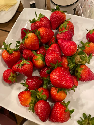 A high-angle, medium shot captures a close-up view of a white, square plate filled with fresh strawberries. The strawberries are a vibrant red, with some showing hints of orange and yellow, indicating varying degrees of ripeness. Their green stems and leaves are visible, adding a natural touch to the arrangement. The plate is positioned on a wooden surface, suggesting it might be on a table or countertop. In the background, out of focus, are glimpses of what appear to be other food items and kitchenware, including slices of cheese on a white plate and a black and white mug. The lighting is bright, highlighting the textures and colors of the fruit. The scene is static and does not depict any actions or interactions. The location of Englewood, United States, is not discernible from the visual information provided in the image.