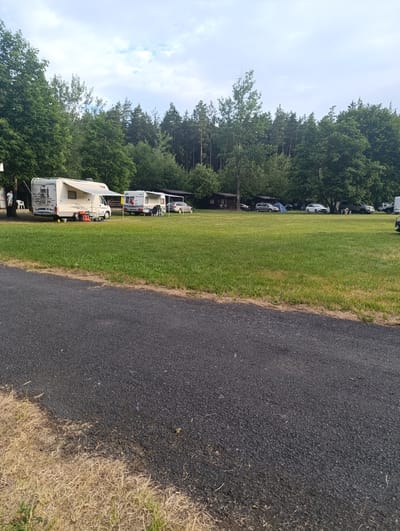 This is a photograph taken from a slightly elevated perspective, looking across a grassy field towards a wooded area. In the foreground, a dark asphalt path runs diagonally from the bottom left to the upper right corner, bordered by dry, light-colored grass. The middle ground features a well-maintained green lawn with several recreational vehicles (RVs) and cars parked.
On the left side of the field, a white RV with an extended awning is prominent, with a person standing near it and several white chairs. To its right, another, smaller white RV is parked, also with an awning extended, and a couple of chairs and a table setup. Further into the field, various cars, including silver and white sedans, are parked amongst the grass. A blue tent is visible between two of the cars.
The background is dominated by a dense line of trees, forming a natural boundary to the field. The sky above is partly cloudy with a mix of white and grey clouds. The scene appears to be a campground or a similar recreational area, possibly located in or near the city of Babylon, Czechia, given the context provided. The overall impression is one of a peaceful outdoor setting, likely during daylight hours.