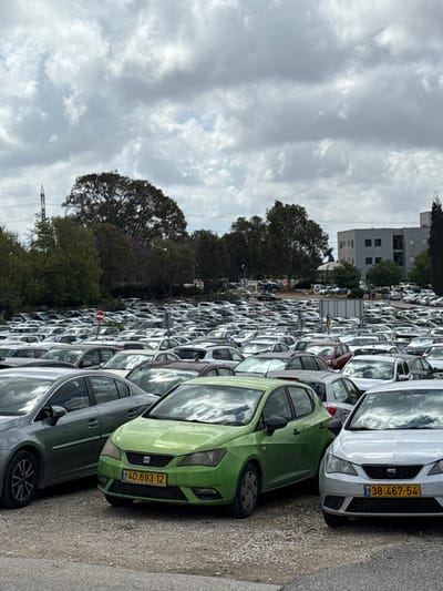The image shows a large parking lot in Kfar Monash, Israel, located at 32.342 latitude and 34.911 longitude. The parking lot is filled with cars, mostly silver or white in color, with a few green and black cars mixed in. The cars are all parked in rows, and many of them have their windows down. In the foreground, a green car with a yellow license plate that reads "40 693 12" is parked next to a silver car with a yellow license plate that reads "38 467 54." The sky is partly cloudy, with some white clouds visible against a light blue sky. In the background, a row of trees and a building with a flat roof are visible.  The image was taken from a slightly elevated position, looking down at the parking lot.  A stop sign is visible to the left.