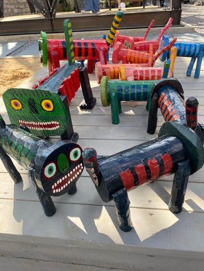 The image shows a collection of colorful, wooden, animal-shaped sculptures placed on a wooden deck. The sculptures are predominantly cylindrical with four legs and decorative elements painted on them. Some of the sculptures are red with black stripes, others are green with yellow and pink eyes and white teeth, and one is blue with black stripes. The background suggests an outdoor setting with sand and a stone wall visible, possibly near some industrial-looking metal structures. A person's legs wearing blue jeans and pink shoes are partially visible in the upper background, walking away from the camera. The scene appears to be in Staraya Voskresenovka, Russia.