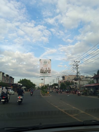Captured from within a vehicle, the scene portrays a street in Payameuneng, Indonesia. The road ahead accommodates a stream of motorbikes, while pedestrians navigate the sidewalks and shopfronts lining the street. A prominent billboard displays an advertisement for "Idul Adha" and features images of public figures, including H. Ruslan M. Daud, S.E., M.A.P., identified as a member of the Komisi V DPR-RI PPKB and Ketua DPW PKB Aceh. Overhead, a patchwork of blue sky and clouds serves as a backdrop to the scene, with electrical wires and poles punctuating the skyline. The street itself is marked with painted lines, indicating traffic lanes.