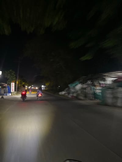 The image shows a street scene at night in Crewek, Indonesia. Two people on motorcycles are traveling down the road. The motorcycle in the foreground has its headlights on, illuminating the road ahead. The motorcycle in the background has its taillight on. There are trees on either side of the road, and a building on the right side of the image. The image is blurry, suggesting that it was taken from a moving vehicle.