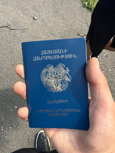 An individual's hand is holding an Armenian passport. The passport is dark blue with gold lettering and the Armenian coat of arms embossed on the cover. The text on the passport is in Armenian and English, reading "REPUBLIC OF ARMENIA PASSPORT". The background consists of a cracked asphalt surface, with a patch of green grass and small white flowers visible in the upper right corner. The image is taken from a slightly elevated perspective, looking down at the passport and the hand holding it.