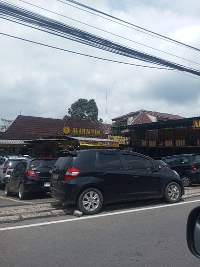 The image depicts a daytime street view of a commercial area, identified as being in Chori, Nigeria, although prominent signage for "ALAM SUNDA," a known Indonesian Sundanese restaurant chain, is clearly visible, along with other Indonesian text on banners.
The scene is captured from a low vantage point, possibly from within a vehicle, with a portion of a side mirror visible in the bottom right. The top third of the frame shows an overcast sky with numerous black power or communication lines stretching horizontally across.
In the midground, a row of vehicles is parked parallel to a concrete curb that separates a paved area from an asphalt road. The road has a clear solid white line. The most prominent vehicle is a black Honda Jazz/Fit hatchback with tinted windows, a rear spoiler, and silver multi-spoke alloy wheels. A small rock or debris is visible against the curb near its rear-right wheel. To its left, another black hatchback, possibly a Honda Brio, is partially visible, showing a license plate that begins with "B 28XX XX". Other parked vehicles are partially obscured further left.
Behind the parked cars stands a restaurant building, likely "Alam Sunda," characterized by its extensive brown-tiled, multi-pitched roof. A large golden "ALAM SUNDA" sign is centrally displayed on the building's facade. Below it, open-air dining areas with wooden railings are visible, where several indistinct figures, presumably patrons, are seated at tables. Banners with Indonesian text, such as "NIKMATI SENSASI IKAN..." (Enjoy the sensation of fish...) and "ALAM KINI," hang from the restaurant's structure.
In the background, more green foliage and trees are visible, along with another building featuring a reddish-tiled roof on the right. A tall, thin, grey mast or antenna rises above the structures in the central background.