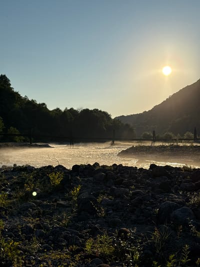 The image depicts a serene riverside scene at what appears to be sunset or sunrise, given the low angle of the sun casting a warm glow. The sun itself is a bright, diffused orb in the upper left portion of the frame, illuminating the sky with a pale yellow hue. The scene is oriented vertically, suggesting the photo was taken in landscape mode and then rotated.
The main subjects include a body of water, likely a river or stream, reflecting the sunlight and appearing shimmering and somewhat misty. Along the far bank of the water, a line of lush green trees and foliage forms a dense forest that climbs a gentle slope towards a silhouetted mountain range in the background. The presence of a few vertical posts or markers in the water suggests a shallow area, a weir, or a shallow crossing.
The foreground is dominated by a rocky shoreline, with large, dark stones and scattered green vegetation. The texture of the rocks is rough and uneven, with some smaller plants growing in crevices. The sunlight highlights the edges and surfaces of these rocks, creating a strong contrast with the shaded areas.
The overall mood is peaceful and tranquil, evoking a sense of natural beauty and quietude. The light is soft and diffused, contributing to the calm atmosphere. There is no visible text in the image. The location is identified as Abgara, Abkhazia. The weather appears clear, with the sun breaking through. No people or specific objects beyond the natural landscape elements and the suggestive posts in the water are identifiable.
