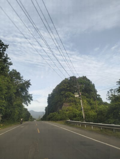 The image captures a scenic view of a road winding through a lush, green landscape in Babah Dua, Indonesia. The road, paved and marked with a yellow dividing line, recedes into the distance, bordered by dense foliage on either side. A single motorcyclist, wearing a helmet and white attire, is visible on the left side of the road, appearing to be pulled over.

The scene is characterized by a sense of tranquility, possibly during the early morning or late afternoon, given the soft, diffused light and the cloudy sky. The sky is a blend of pale blue and white, with streaks of clouds creating a dynamic texture. Overhead, a set of parallel power lines stretch across the frame, casting faint shadows on the sky.

Dominating the right side of the road is a large, verdant hill or rock formation, heavily covered in trees. A utility pole with electrical equipment stands prominently against the green backdrop. In the distance, rolling hills can be seen, adding depth to the landscape. The overall impression is one of a peaceful rural setting, with nature taking center stage alongside the man-made road and power infrastructure. There is no visible text in the image.
