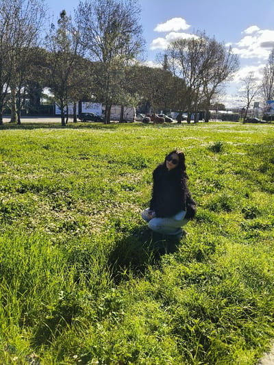The image displays an outdoor scene in Riccione, Italy, under clear, sunny conditions. A sole female individual is crouched in the foreground, situated slightly to the right of the frame on a vibrant green grassy field. She is wearing a dark, fuzzy jacket, light blue jeans, and dark sunglasses, looking directly towards the camera with a subtle expression. Her shadow is cast prominently on the grass to her immediate left. The grass contains numerous small white flowers. In the midground, a line of deciduous trees, mostly bare of leaves, runs horizontally, partially obscuring a paved road. Behind these trees and along the road, several vehicles are visible, including a black car, a white recreational vehicle (RV) or camper, and an orange pickup truck. Beyond the road and vehicles, commercial billboards or signs are present, with indistinct structures or buildings visible further in the background. The sky above is blue with scattered white cumulus clouds.